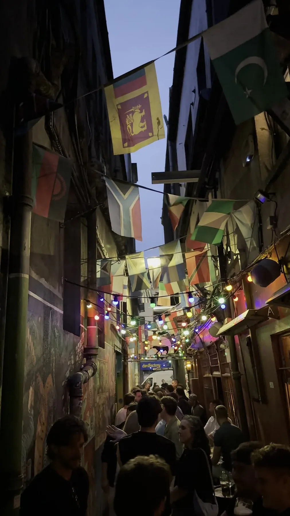 Mutton Lane at dusk with coloured lights, flags, mural and people outside the pub